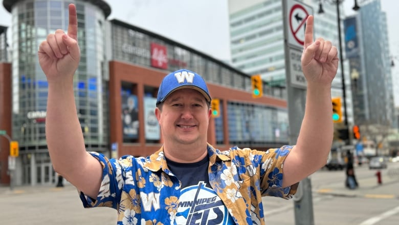 A man in a baseball hat holds up two index fingers in a No. 1 celebration and stands on a street corner.