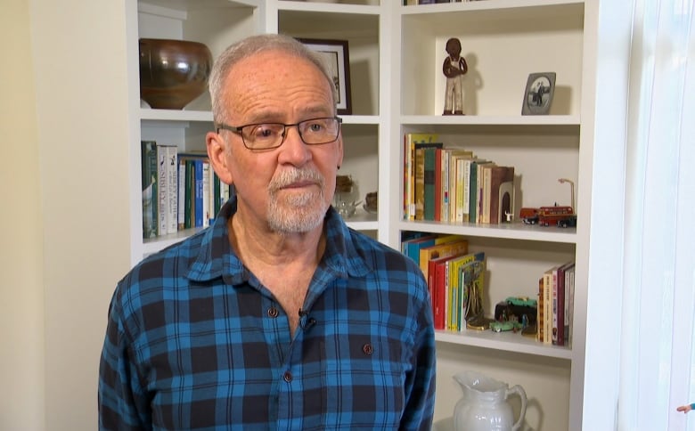 A man with white hair wears glasses and a plaid blue shirt. He stands in front of a white shelving unit with books and photos on display. 