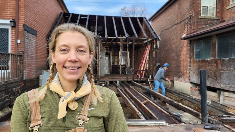 Woman in front of the frame of a house. A man with a hard hat is in the background.