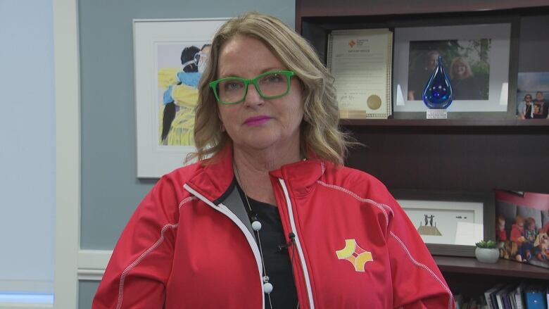 A woman with blonde hair and green glasses sits down and looks at a reporter. She is wearing a red jacket.