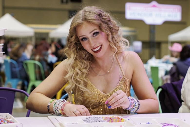 In a slip sequin dress, a Taylor Swift look-alike sits at a table, making friendship bracelets and smiling for the camera.