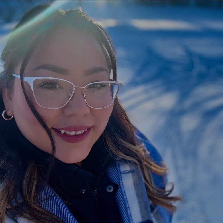 A young girl is posing outside while wearing glasses and hair half up, with snow in the background.