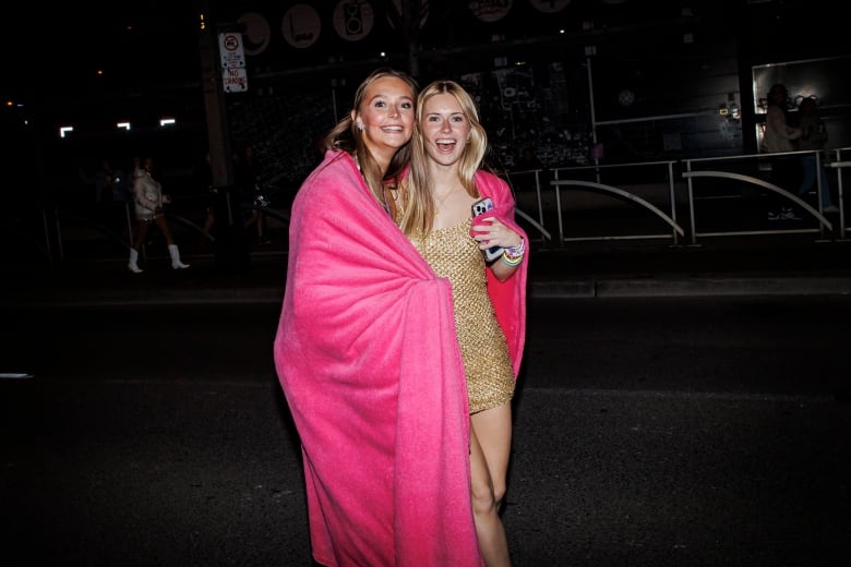 Smiling, two young women in tank dresses, wrapping themselves in a pink blanket, as they wait in a lineup to get inside Rogers Centre stadium for the Taylor Swift Eras Tour concert.