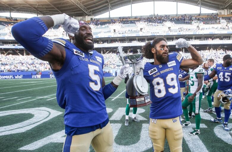 Two players in blue and gold flex with one arm while holding a trophy in the shape of a banjo with their other arms.