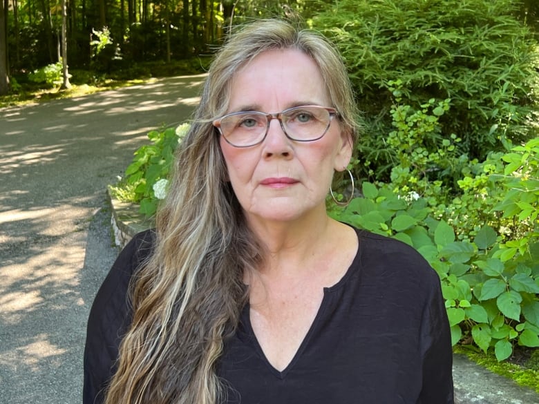 A woman with long blonde hair and glasses wearing a black top looks directly at the camera with a neutral facial expression.