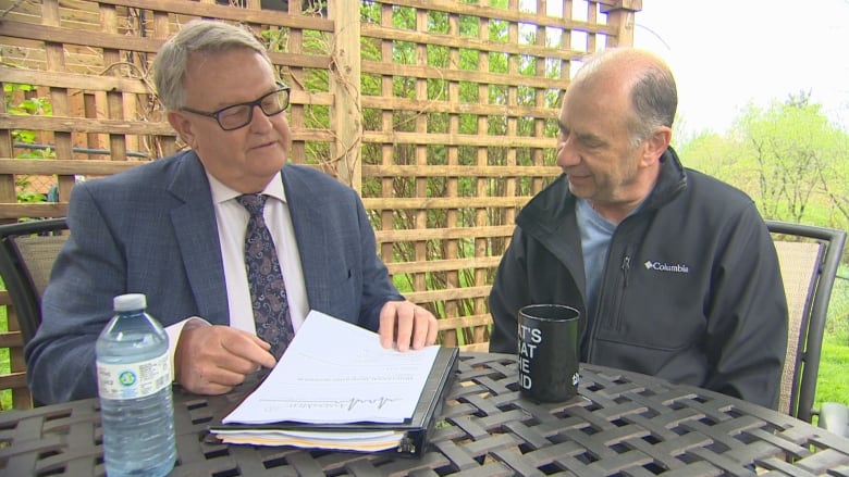 Two men sit at an outdoor table looking through some documents together. The one on the left wears glasses, a blue suit, and a tie, and the man on the right is wearing a blue v-neck shirt with a black jacket over it.