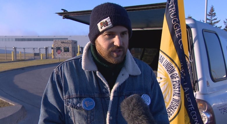 A man wearing a black beanie and jean jacket speaks into a microphone, with a flag and vehicle behind him.