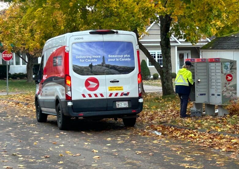 A Canada post van parked on the side of the paved road, while a postal worker in a bright yellow jacket fills post office lock-boxes. The season is fall. 