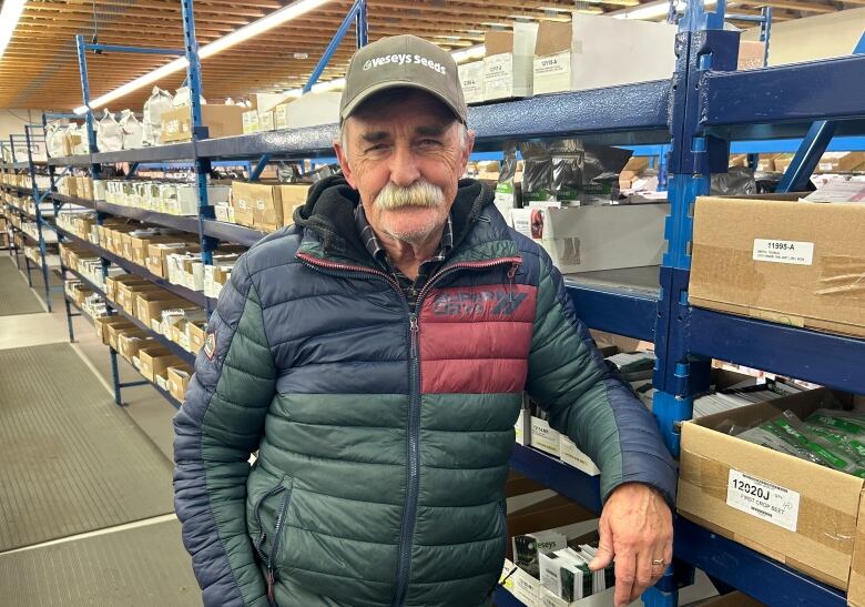 A man in a forest green and navy puffy jacket and ballcap, gray hair and a bushy gray mustache, looks at camera as he leans against rows of product in a warehouse. 