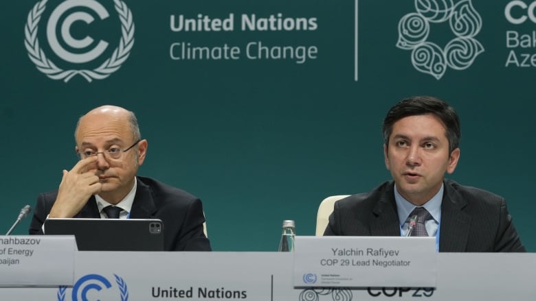 Two men sit at a panel in front of a teal backdrop that says United Nations Climate Change. The man on the left is older and scratching his nose, looking bored. The man on the right appears to be in the middle of talking. 