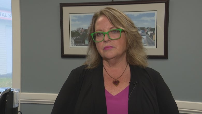 A woman with blonde hair and green glasses sits down and looks at a reporter.