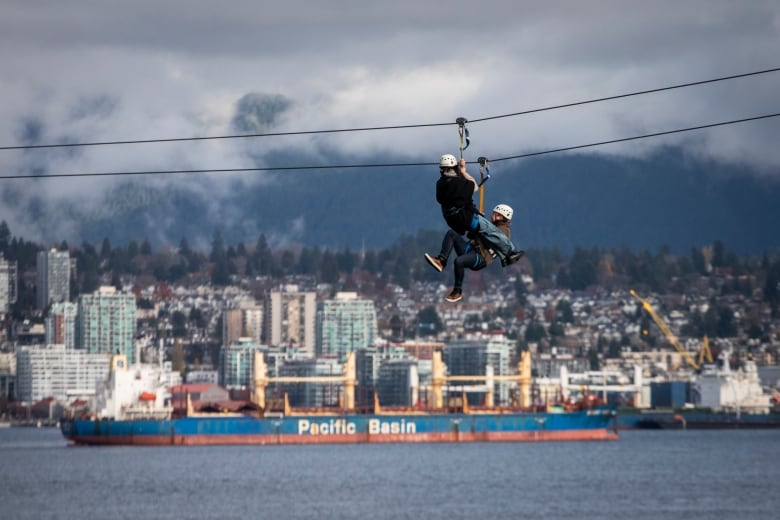 Two people in helmets slide down a zip line over water with a city skyline in the background.