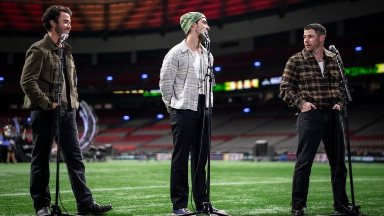 Three brothers stand next to each other on a football field while speaking into microphones during a press conference. 