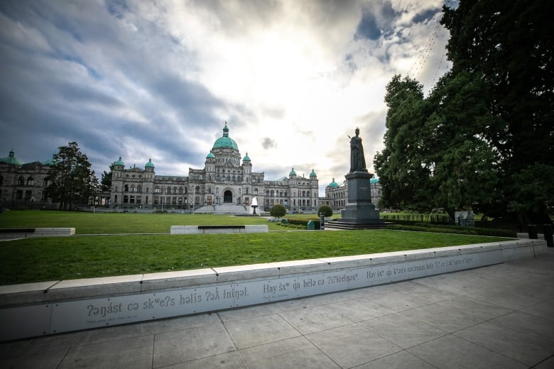 A Legislature building looms in the background on a cloudy day, with a low wall displaying an Indigenous language in the forefront.