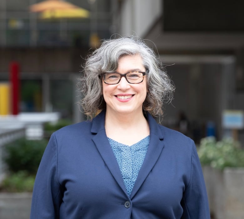 A woman with graying, shoulder length wavy dark hair wearing a cobalt blue blazer and blouse smiles in a head and shoulders portrait. The background is blurred but appears to be an exterior of an institutional building.