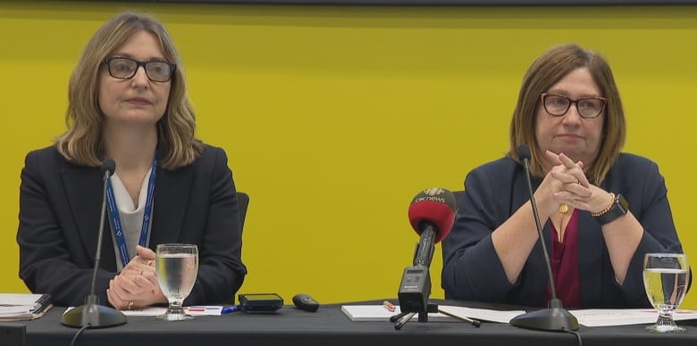 Two women with black blazers sit behind table with microphones