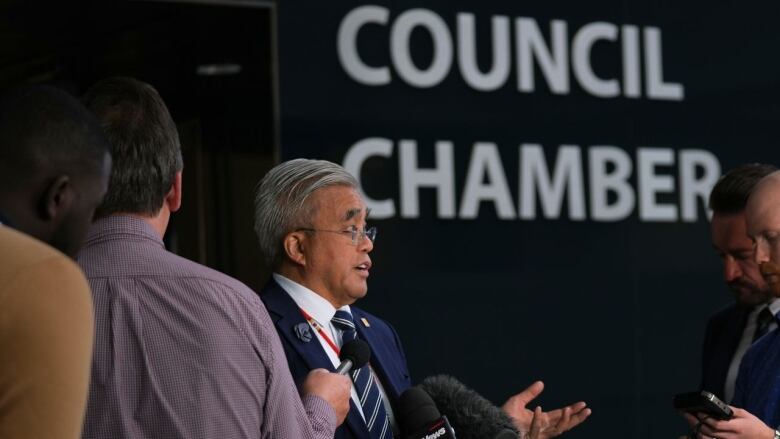 a man speaks to reporters in front of a sign that reads 