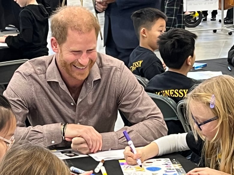 A smiling man sits in a gym full of kids.