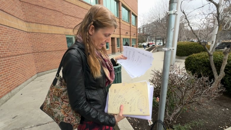 A woman with long red hair in a slim winter jacket examines a folder of legal documents on the courthouse steps in Kelowna, B.C. 