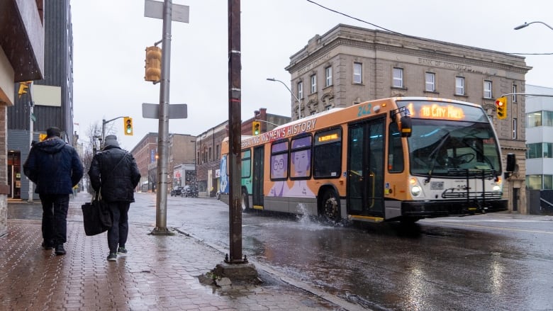 Two people walk on the sidewalk in the rain while a bus passes kicking water up under the tire