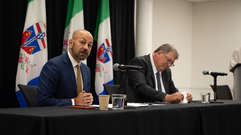 Two men in suits at table in front of flag.