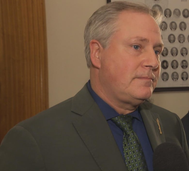 A man in a grey suit stands in a hallway talking to reporters.