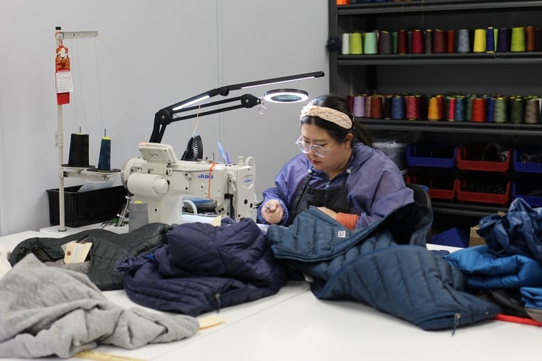 A woman wearing glasses, a white headband, a purple jacket and black apron works at a sewing machine to repair a blue puffer jacket.