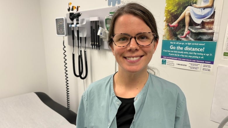 A women wearing glasses and smiling at the camera. She is standing in a doctors office and is wearing a lab coat. 