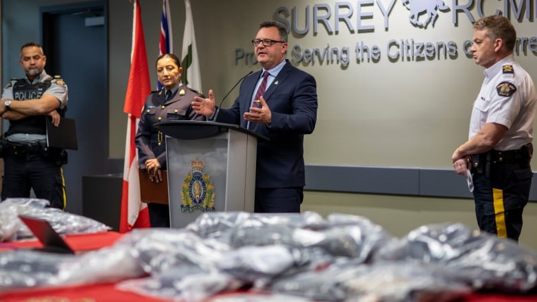 A man in a suit stands at a podium flanked by uniformed RCMP officers in front of a display of bagged drugs sitting on a table covered in red cloth in front of them.