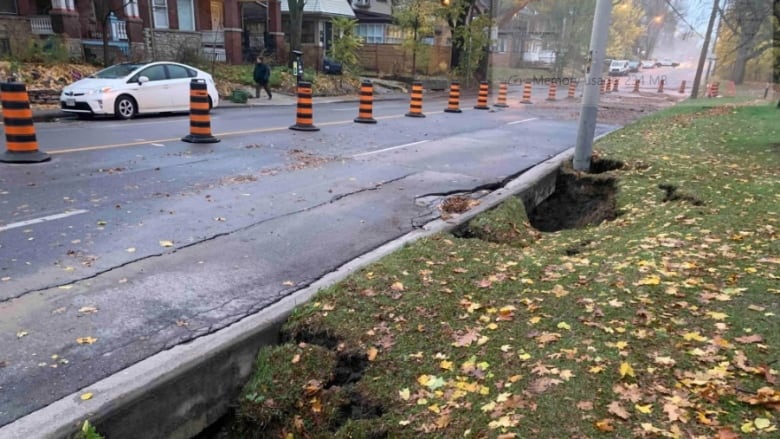 2 Sinkholes on the side of a residential city street in fall. There are pylons closing the road to traffic. It's a wet fall day