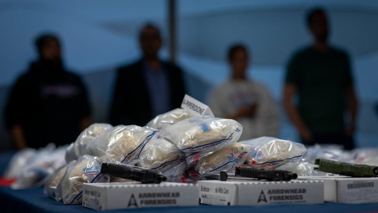 four figures, nearly silhouettes, stand in the background behind a pile of bagged drugs on a table. There are three handguns on white cardboard boxes that say 