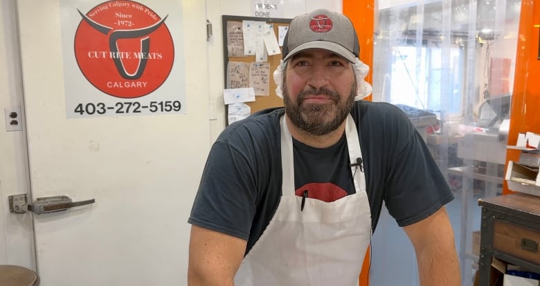 A butcher in a white apron stands behind the counter of his meat shop.