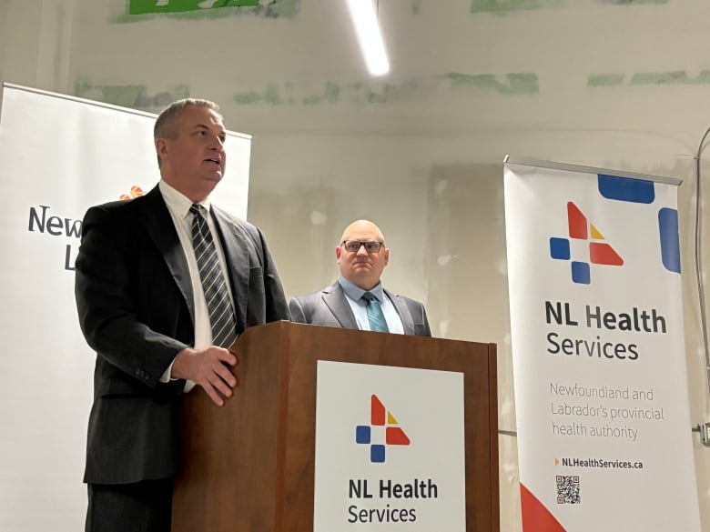 Two men wearing suits stand in an uncompleted room in a hospital. There are banners for Newfoundland and Labrador Health Services, and the man closest to the camera is standing at a podium.