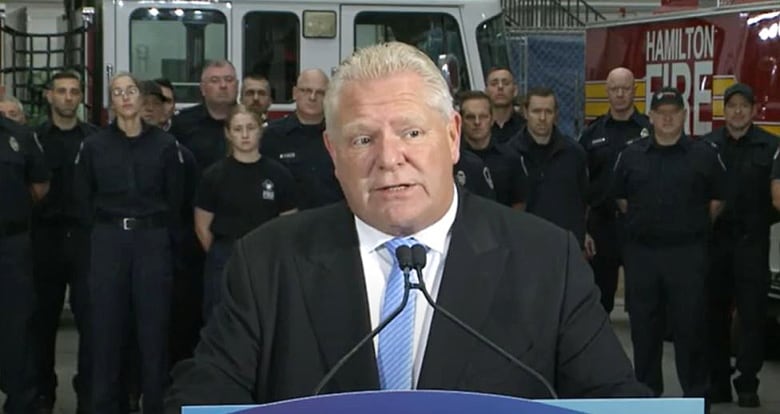 Man standing at podiumn in a firehall with firefighters standing behind him