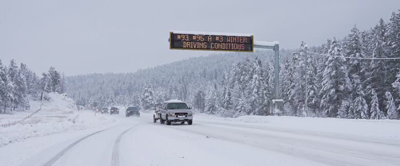 A row of cars and pickup trucks drive past a snowy highway, with a sign reading '#93 #95 A #3 Winter Driving Conditions.'