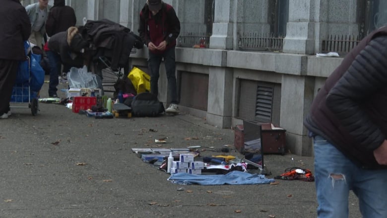 People gather around as a carpet with various small items on it is pictured on a sidewalk.