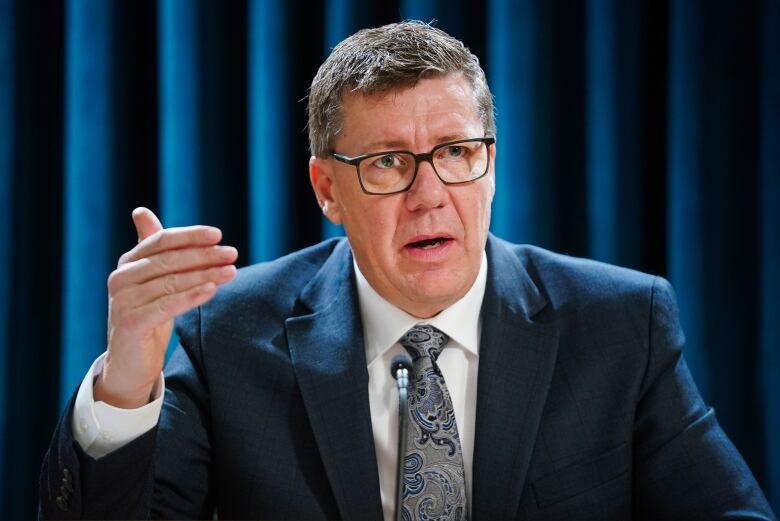 A man with glasses and a suit gestures while speaking.